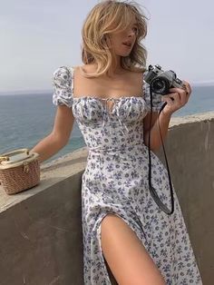 a woman in a dress is holding a camera and leaning against a wall near the ocean