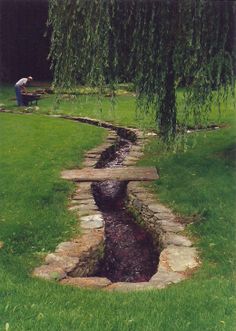 a man sitting on a bench next to a small creek in the grass near a tree