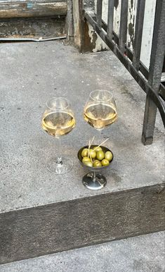 two glasses of wine and some olives in a bowl on the ground next to an iron railing