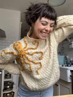 a woman is smiling in the kitchen with her hands on her head and wearing an octopus sweater