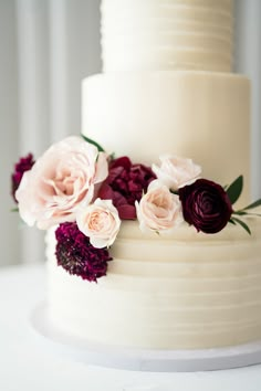 a three tiered white cake with purple and pink flowers on the top, sitting on a table