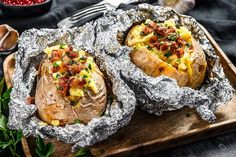 two baked potatoes covered in bacon and cheese on a wooden cutting board with silver foil