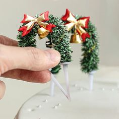 a hand is holding two small christmas wreaths on top of a white cake with gold and red bows