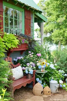 a red bench sitting in front of a green house surrounded by plants and flowers on the ground