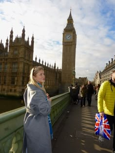 two people standing on a bridge with big ben in the background and other people walking by