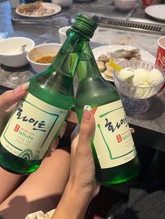 two bottles of beer are being held up by someone's hands at a table full of food