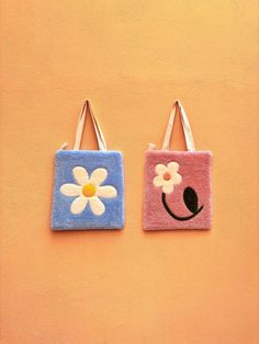 two handbags with flowers on them hanging from hooks against a yellow wall, one has a smiley face and the other has a flower