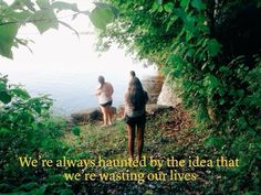 two people standing on the edge of a cliff with trees and water in the background