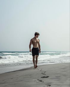 a shirtless man walking on the beach next to the ocean with footprints in the sand