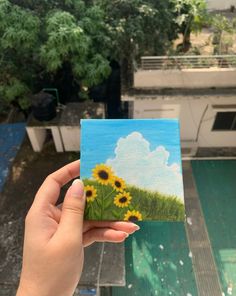 a person holding up a small card with sunflowers painted on it in front of a building