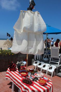 a red and white table topped with food under an umbrella on top of a beach