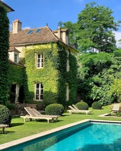 an outdoor swimming pool surrounded by lush green plants and lawn furniture next to a large house