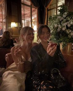 two beautiful women sitting next to each other at a table with wine glasses in their hands