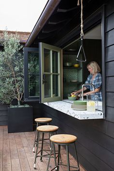 three stools sit on a wooden deck near an outdoor kitchen area with black walls and windows