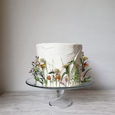 a white cake with flowers on it sitting on top of a glass platter in front of a wall