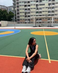 a woman sitting on the ground in front of a basketball court with buildings behind her