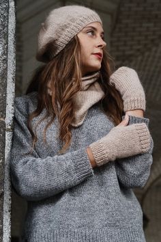 a woman with long hair wearing a gray sweater and mittens is leaning against a wall