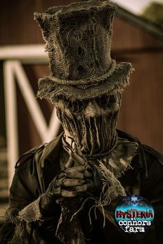 a creepy looking man wearing a hat and holding his hands to his face while standing in front of a barn