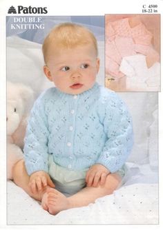 a baby wearing a blue knitted cardigan and white pants sitting next to a teddy bear