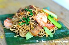 a plate of noodles with shrimp and vegetables on a leafy green plate sitting on a wooden table