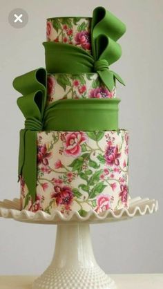 a three tiered cake with green ribbons and flowers on it's side, sitting on top of a white pedestal