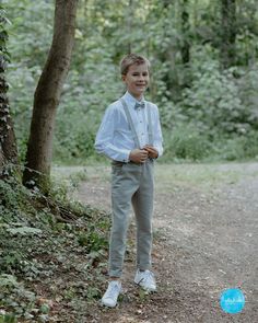 a young boy in a white shirt and tie standing on a dirt road next to trees