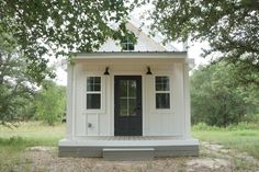 a small white house with black doors and windows on the front porch is surrounded by trees