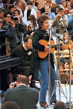 a man holding a guitar in front of a microphone while standing next to a crowd