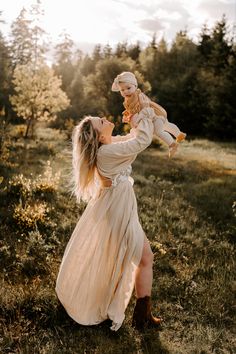 a woman holding a baby in her arms while standing on the grass with trees in the background