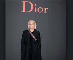 an older woman standing in front of a dior sign wearing a long black coat