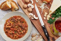 a wooden cutting board topped with pasta and meat next to garlic bread, tomatoes, mushrooms, parmesan cheese and tomato sauce