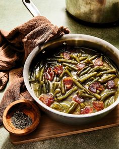 a pot filled with green beans and bacon on top of a cutting board next to a wooden spoon