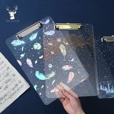 three clear folders with designs on them and music sheets in the background, one being held up by a person's hand