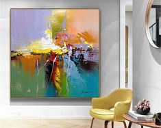 an abstract painting hangs on the wall next to a yellow chair and round table in a white room