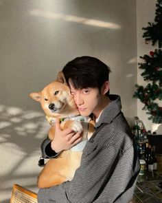 a man holding a dog in his arms while sitting on a chair next to a christmas tree