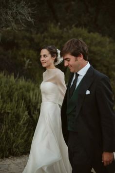 a bride and groom walking down the street together in their wedding attire, with greenery behind them