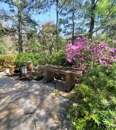 a wooden bench sitting on top of a stone walkway next to trees and bushes with purple flowers in the background