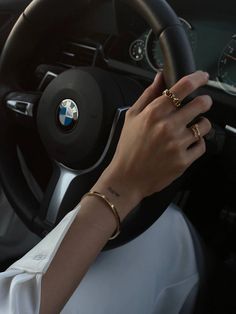 a woman driving a car with her hand on the steering wheel and wearing gold rings