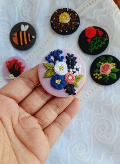 a hand holding a small embroidered brooch with flowers and leaves on it, surrounded by other buttons