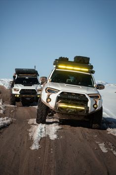 two white trucks driving down a snow covered road in the middle of winter with their lights on