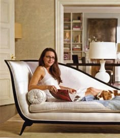 a woman sitting on a white couch with a dog laying next to her and reading a book