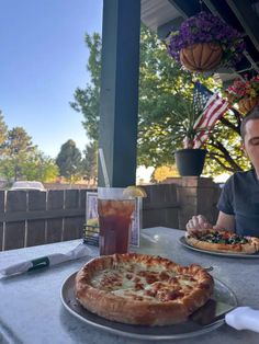 a man sitting at an outdoor table with two pizzas on it and a drink in front of him