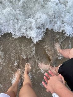 two people sitting on the beach with their feet in the water and one person standing next to them