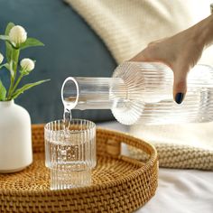 a person pouring water into a glass on top of a wicker tray next to a vase with flowers