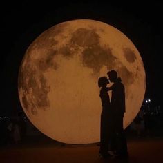 two people standing in front of the moon at night with their arms around each other