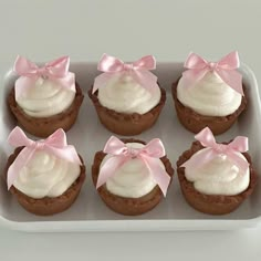 six cupcakes with white frosting and pink bows in a plastic container on a table