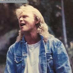 a man with long blonde hair wearing a jean jacket and white t - shirt laughing