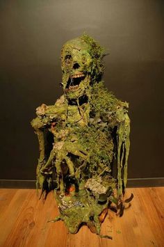 a creepy looking statue made out of moss on a wooden floor in front of a black wall