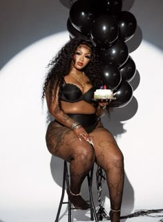 a woman sitting on a chair holding a piece of cake in her hand and balloons behind her