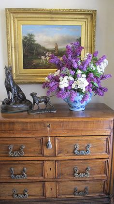 a vase filled with purple and white flowers sitting on top of a dresser next to a painting